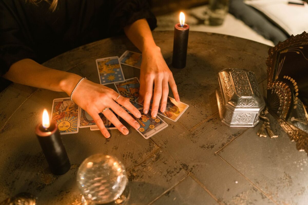 Mãos abrindo poucas cartas sobre a mesa, entre velas e uma bola de cristal