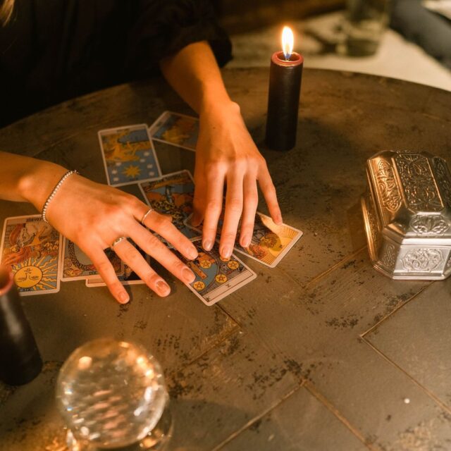 Mãos abrindo poucas cartas sobre a mesa, entre velas e uma bola de cristal